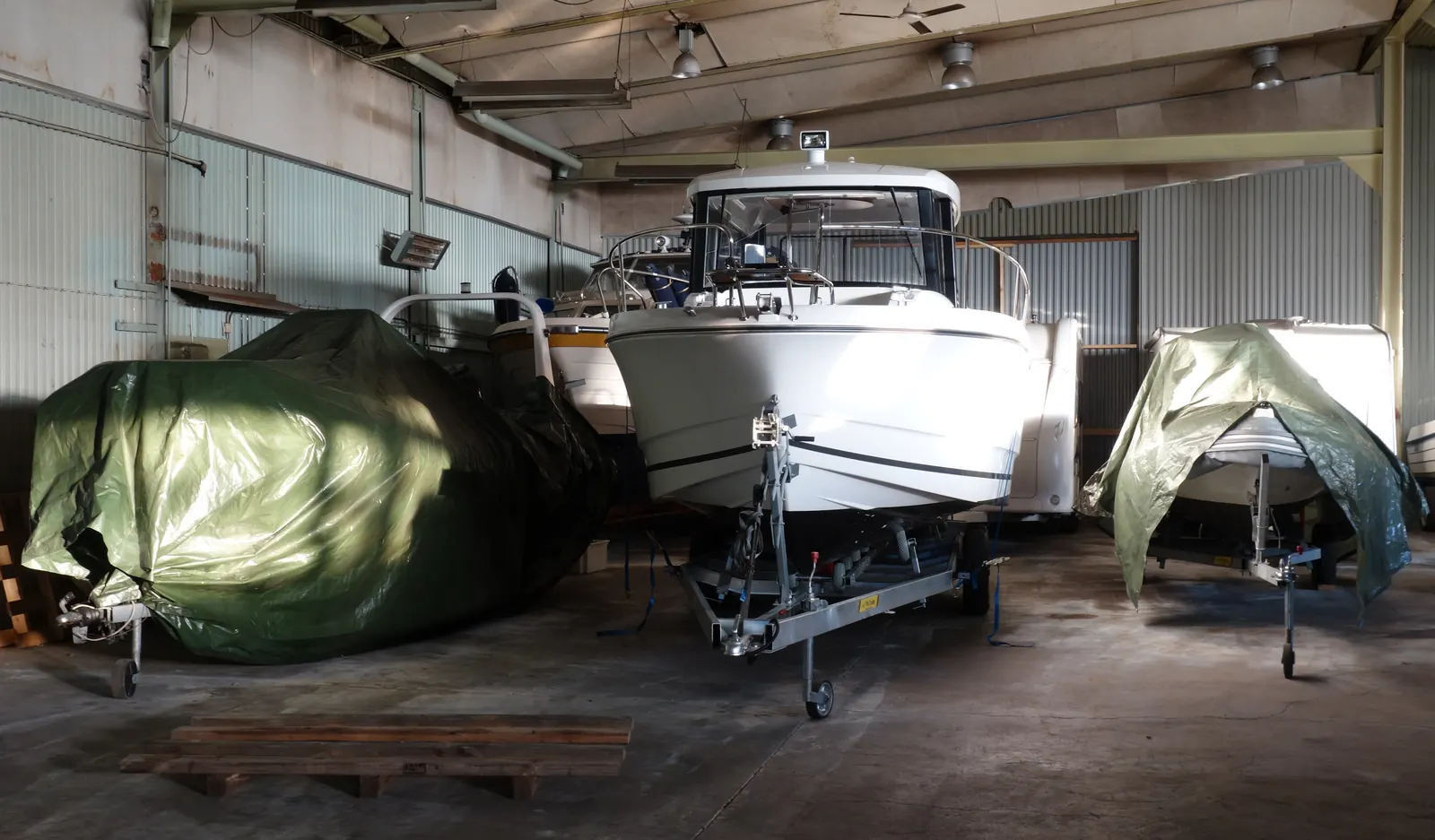 Inside a corrugated-metal storage hall, a white cabin motorboat sits on a galvanized trailer beside a boat wrapped in a green tarp on its own trailer, with more boats behind and a wooden pallet on the concrete floor.