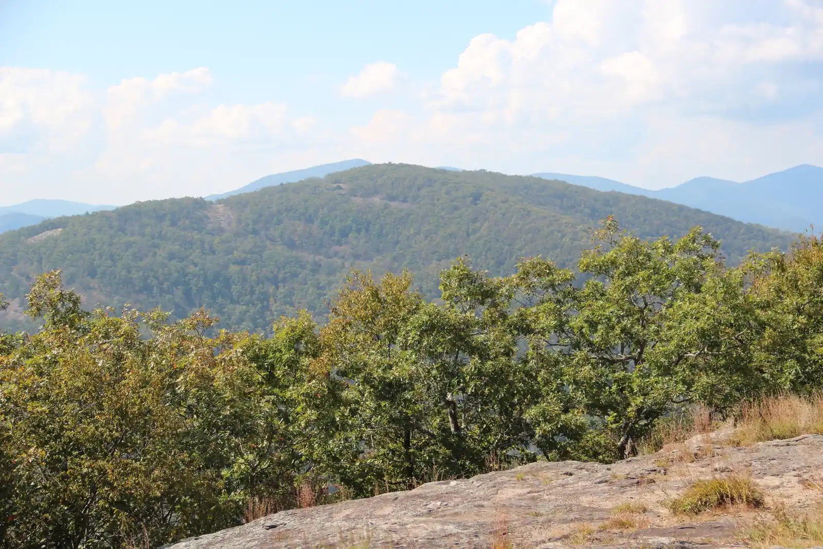 A broad forested mountain, Cowrock Mountain, rises beyond a foreground of leafy oaks and a bare granite outcrop on Wildcat Mountain, with more blue ridges behind under a partly cloudy blue sky in early October.