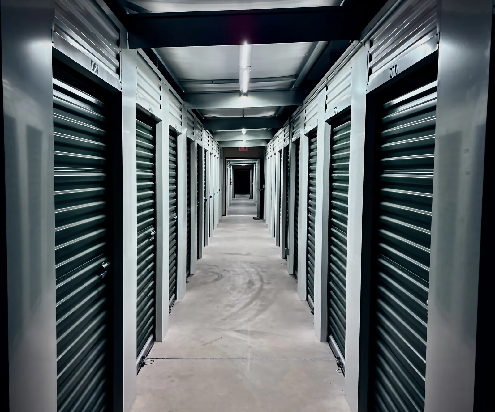 Moody, low-light indoor hallway of climate-controlled storage units, with dark roll-up doors on both sides labeled D67 and D70, steel ceiling beams and a line of lights receding to a red exit sign at the far end. Portrait orientation.