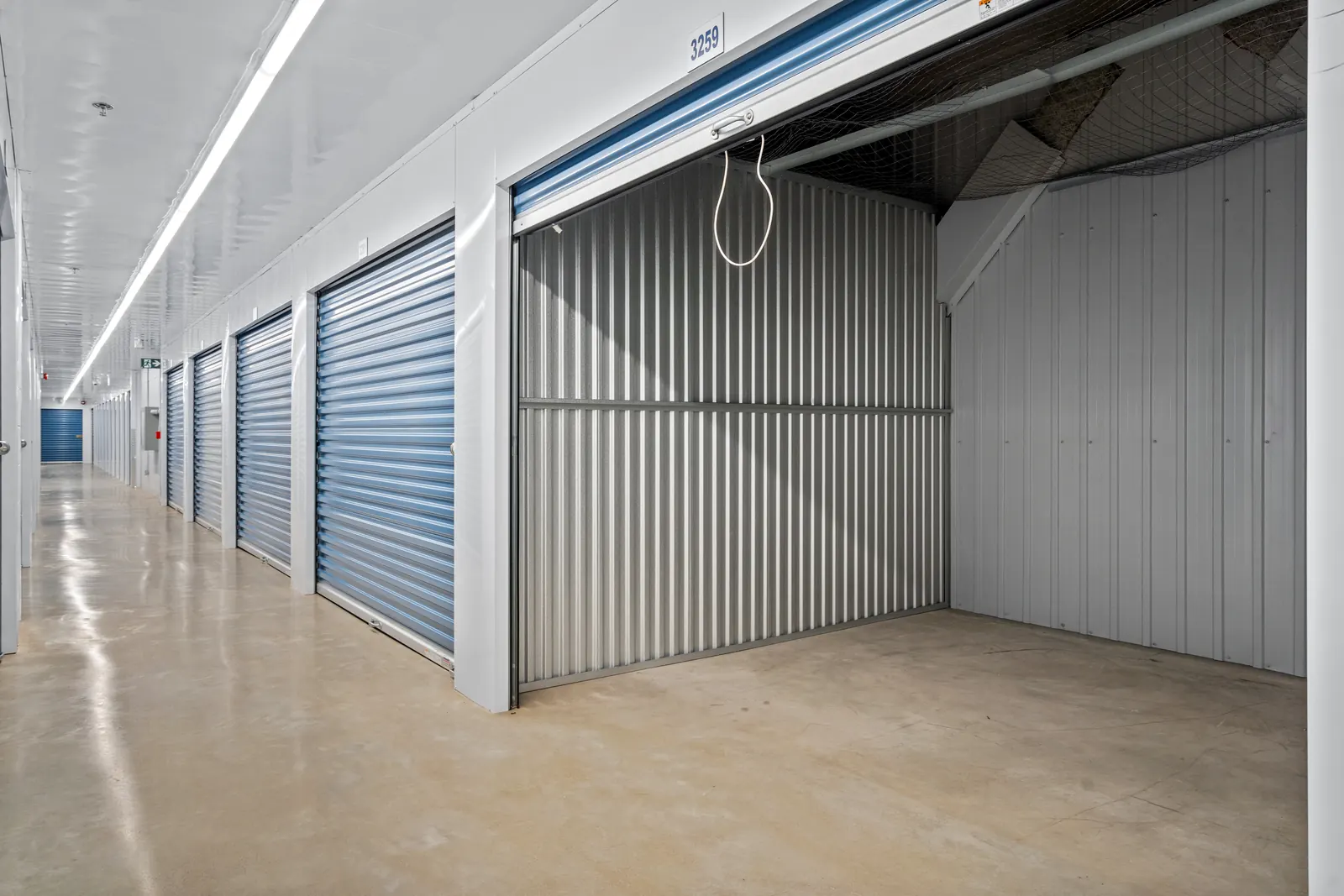 Brightly lit indoor storage corridor with glossy concrete floor, white walls and a row of blue roll-up doors, the nearest raised to show an empty unit with corrugated metal walls; a long ceiling light and a green exit sign run down the hall.