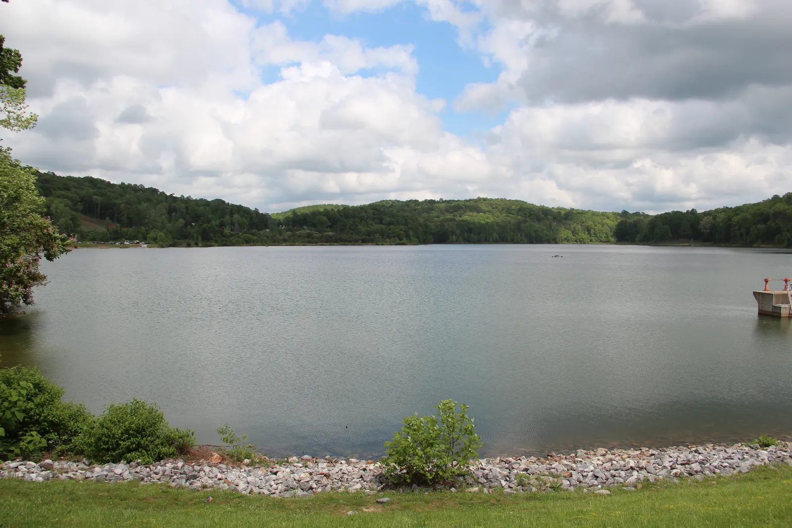 Calm gray-green Lake Zwerner stretches to forested low hills under billowing white clouds and patches of blue sky. A riprap stone shoreline, mowed grass and shrubs fill the foreground, with a small concrete intake structure at the right edge.