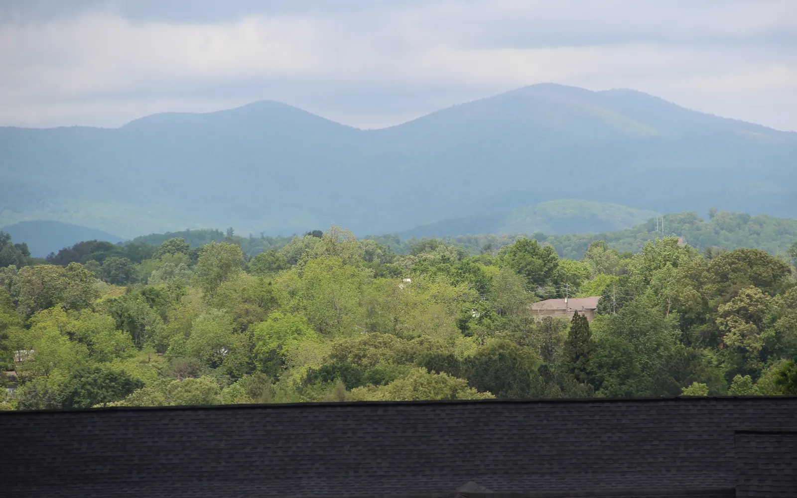 Long Mountain and Gooch Mountain form two blue-green forested peaks beneath an overcast sky, seen from Findley Ridge in Dahlonega above rolling green treetops with a few rooftops. A dark shingled roof edge crosses the bottom of the frame.
