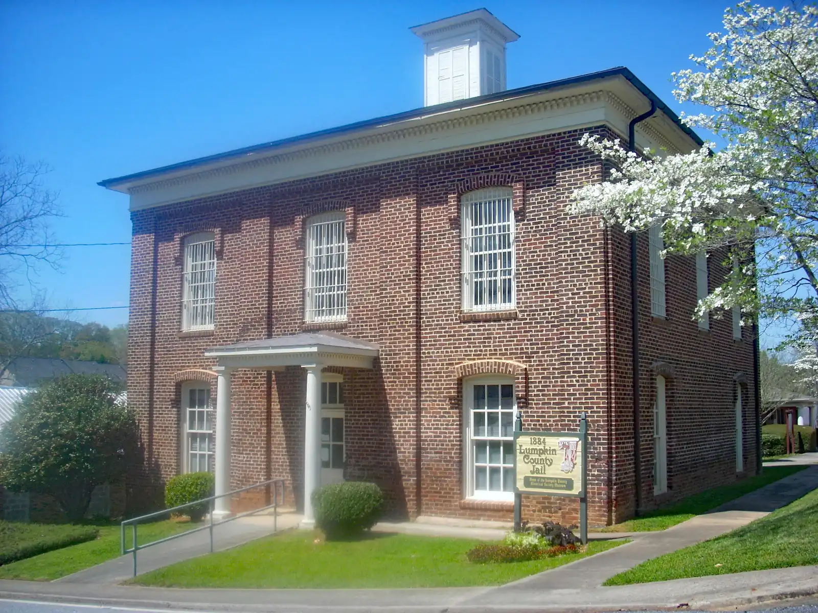 Two-story red-brick 1884 Lumpkin County Jail in Dahlonega with a white cupola, white-columned entry porch and barred upper windows. A white flowering dogwood stands at right, and a wooden sign sits on the green lawn beside a concrete walkway under a clear blue sky.