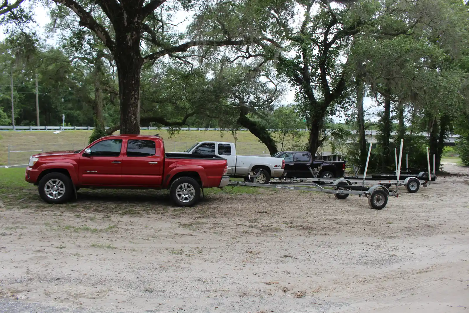 Three pickup trucks, red, silver and black, are parked on sandy ground under large live oaks at a boat-ramp park, each hitched to an empty aluminum boat trailer with white guide posts. A highway guardrail and grassy embankment run behind them.