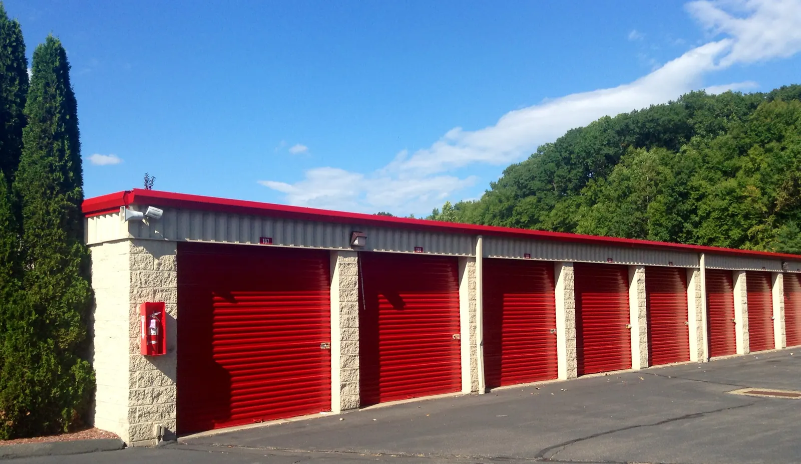 Row of bright red roll-up doors on a single-story self-storage building with split-face block pillars, red roof trim, a security camera and a fire extinguisher in a red cabinet, beside tall evergreen shrubs under a blue sky.