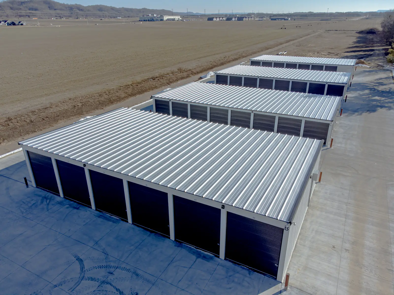 Elevated drone view of four long self-storage buildings with silver standing-seam metal roofs and rows of dark roll-up doors along a concrete drive, beside a bare brown field with a few patches of snow.