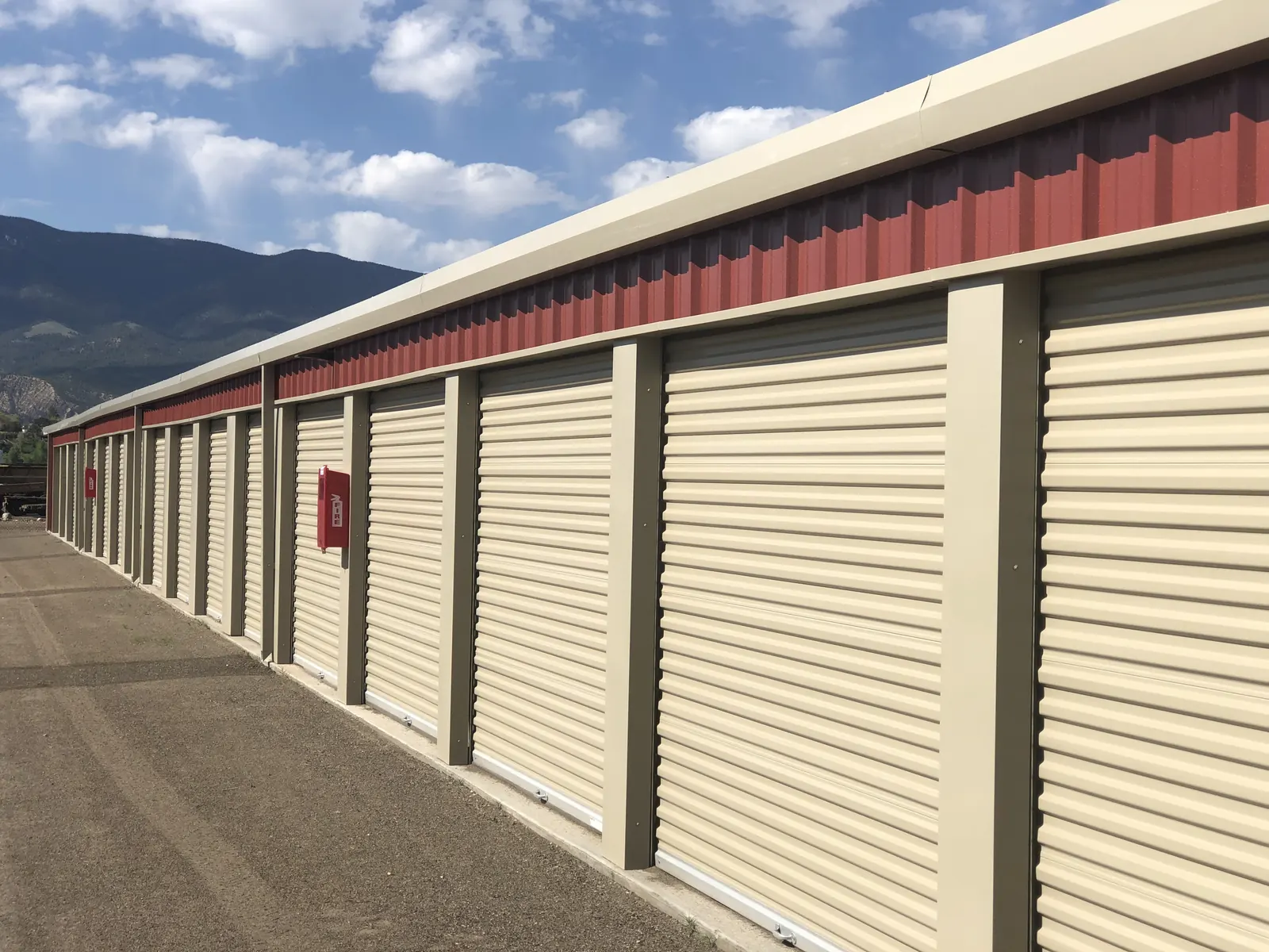 Close angled view of a long row of cream roll-up storage doors set between taupe pillars, under maroon metal siding, with red 'FIRE' extinguisher boxes on the posts and dark mountain slopes in the background.