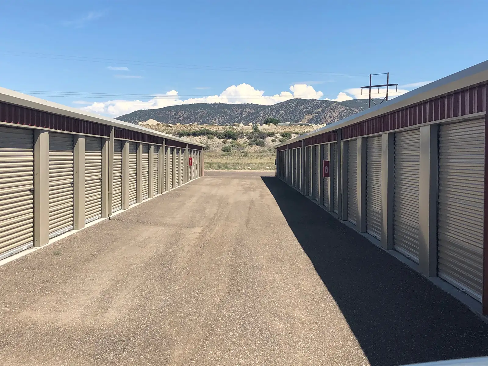 A gravel drive aisle runs between two rows of beige roll-up storage doors with maroon upper siding toward a scrubby hillside, boulders and a forested mountain ridge under a blue sky with white clouds.